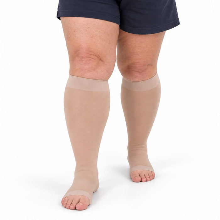 Person med beige kompresjonsstrømper for leggene, i stor størrelse og med åpen tå, avbildet fra leggen og ned mot en hvit bakgrunn.