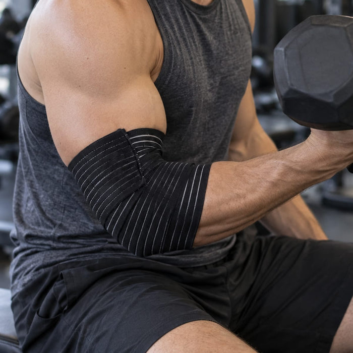 En mann på et treningssenter med elastisk albuebeskyttelse løfter en manual, iført en mørk singlet og shorts.